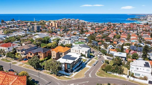 A high-angle aerial view of a dense coastal suburban neighbourhood, showing detached homes, apartment buildings, curved residential streets, and a prominent modern flat-roof house in the foreground. The ocean, headland, and beachfront development are visible beyond the urban area under clear blue skies.
