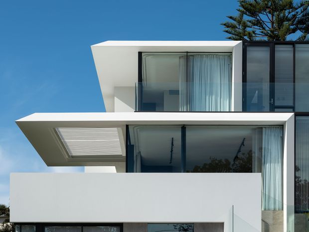 Contemporary multi-level residential facade with crisp white cubic volumes, deep cantilevered slabs, extensive floor-to-ceiling glazing, dark window frames, and a louvred roof opening. The clean geometric composition is set against a clear blue sky, with a tall tree visible behind the building.