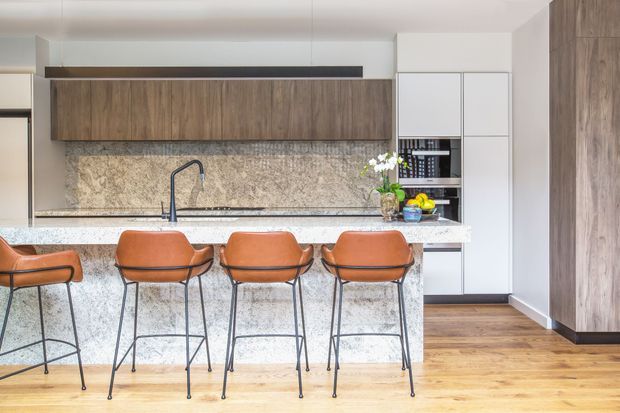 A bright contemporary kitchen with a long island and four rust-colored bar stools. The space features warm wood cabinetry and flooring, white built-in cabinetry, a pale stone island benchtop, a heavily patterned stone backsplash, and a dark gooseneck tap. Full-height wood cabinetry is visible on the right, with open shelving and appliances integrated into the rear joinery.