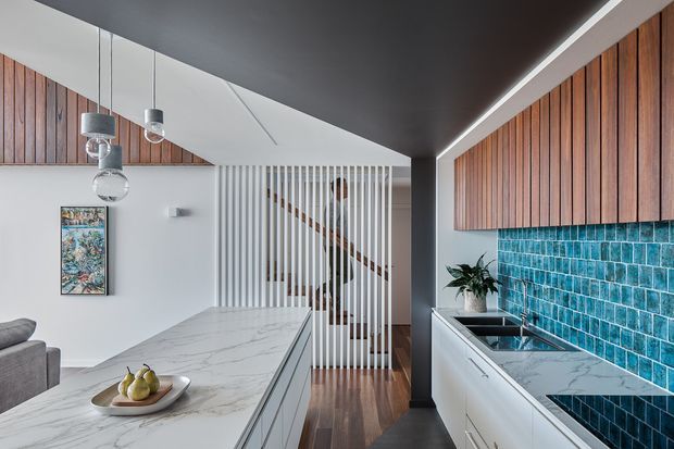 Contemporary open-plan kitchen with a large white marble-look island, white cabinetry, and a vivid turquoise-blue tiled splashback. Vertical timber wall panelling and cabinetry add warmth, while a slatted timber screen partially separates the kitchen from the staircase and adjacent living area. Pendant lights hang above the island, with dark ceiling and structural elements creating contrast.