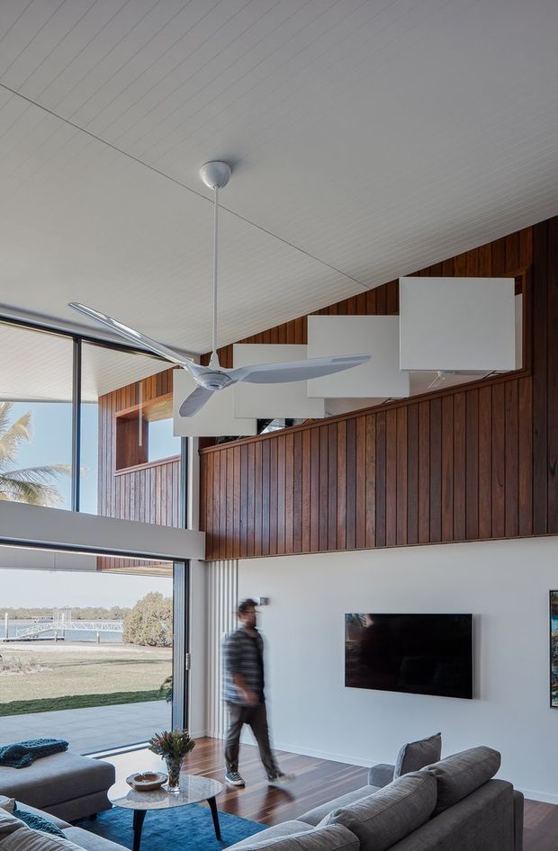 Bright contemporary living room with a dramatic double-height sloped ceiling, exposed dark timber wall panelling, white acoustic ceiling boards, large floor-to-ceiling glazing, and a suspended modern ceiling fan. A grey sectional sofa, round coffee table, television, and minimal furnishings occupy the lower level, with a mezzanine-like upper wall zone and large white acoustic panels. The glazing opens to a landscaped waterfront or riverside setting.