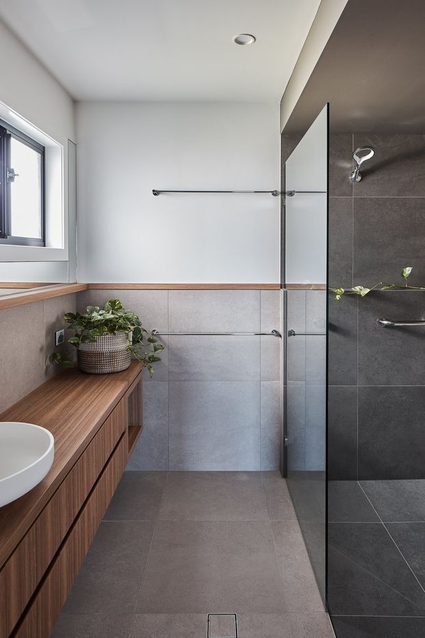 Contemporary bathroom with a walk-in glass shower, wall-mounted brushed-metal towel rails, grey large-format tiled walls and floor, and a warm timber floating vanity with a white vessel basin. A high window brings in natural light, while a potted plant adds greenery.
