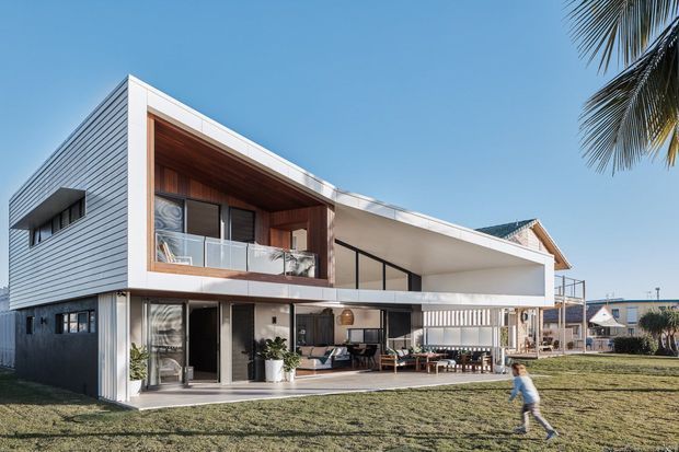 A contemporary two-storey coastal-looking residence viewed from the front garden, featuring crisp white horizontal cladding, a dramatic projecting upper-level form, recessed timber-lined balcony and soffit, extensive glazing, and open-plan indoor-outdoor living areas. The house sits in a suburban setting beneath a clear blue sky, with a lawn and palm visible in the foreground.