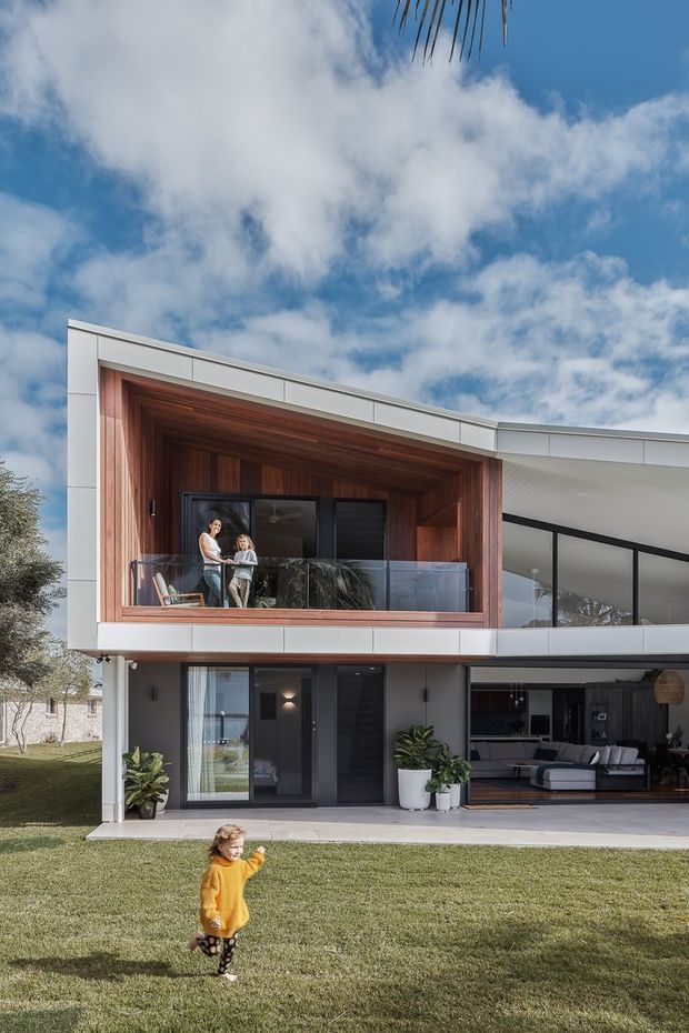 Exterior view of a contemporary two-storey residence with a curved roofline and extensive glazing. The upper level features a deep timber-lined balcony or entertaining recess with a glass balustrade, while the ground floor opens toward a covered outdoor area and visible living room. White panel cladding, dark window frames, timber detailing, landscaped lawn, and a bright partly cloudy sky define the scene.