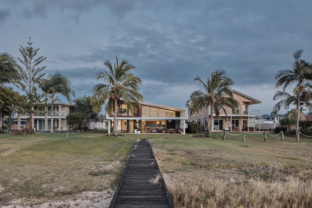 Wide exterior view of a contemporary coastal house set behind a sandy, grassy foreground. A narrow timber boardwalk leads directly toward the home, which features broad glazing, open-plan ground-level spaces, pale rendered walls, and a low pitched roof. Palm trees and neighbouring coastal-style residences frame the property beneath an overcast sky.