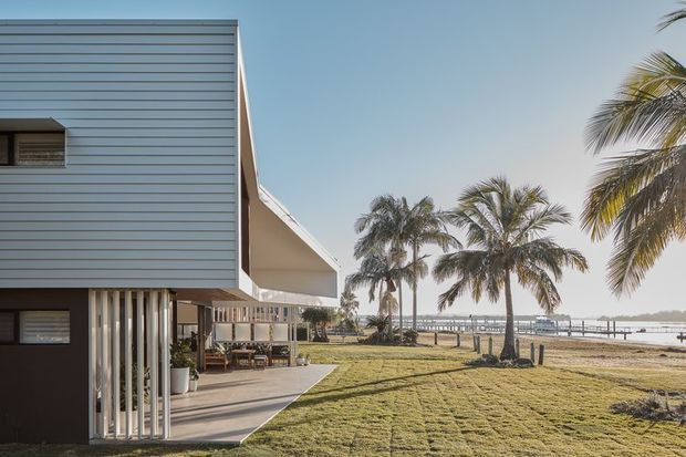 Exterior view of a modern waterfront house with a projecting upper volume clad in horizontal pale grey weatherboards, slender vertical supports, and a covered outdoor living area opening onto a broad lawn. Palm trees and a pier-lined body of water create a tropical coastal setting.