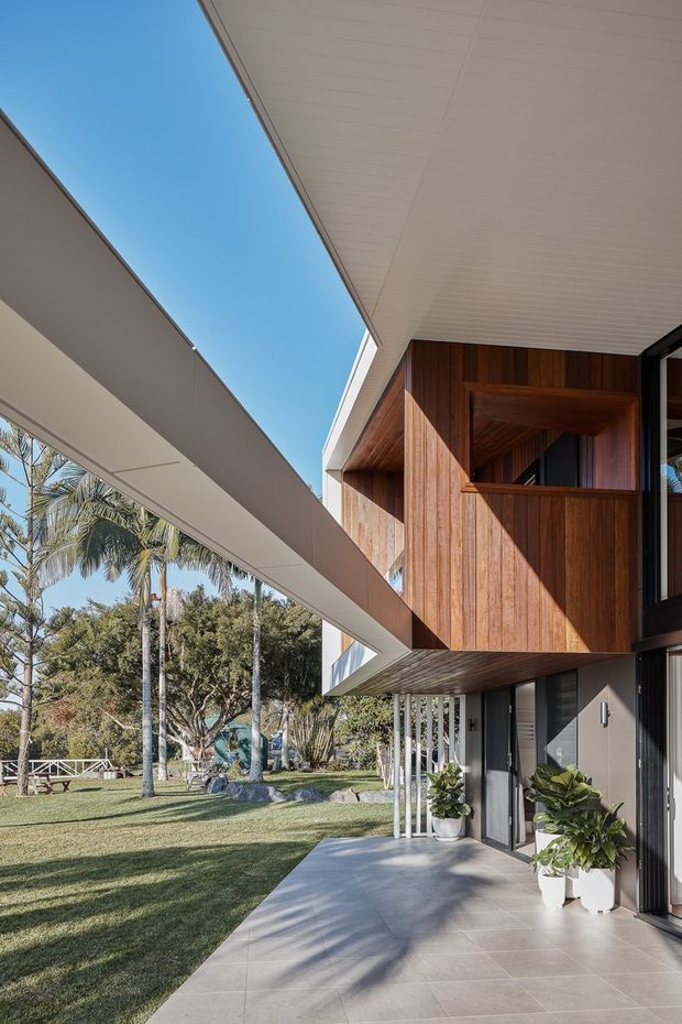Contemporary two-storey residential exterior viewed from a covered patio or walkway. The house features strong cantilevered forms, a broad white-lined soffit, warm vertical timber cladding, dark-framed glazing, and a tiled outdoor terrace. A landscaped garden with palms surrounds the building, and the open sky and deep roof overhang create a bright indoor-outdoor connection.