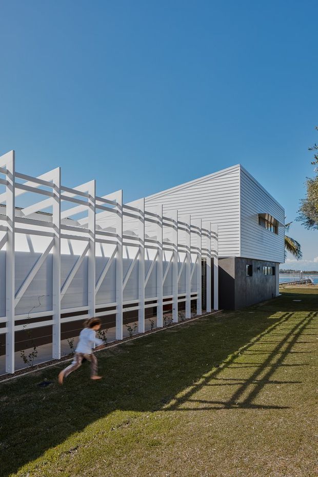 Exterior architectural photograph of a contemporary, low-rise building beside a waterfront. The structure combines a large horizontally lined light-grey upper volume with a darker lower section and a prominent repetitive white structural screen or brise-soleil along the side. Grass surrounds the building, with strong afternoon shadows, clear blue sky, and glimpses of water beyond.
