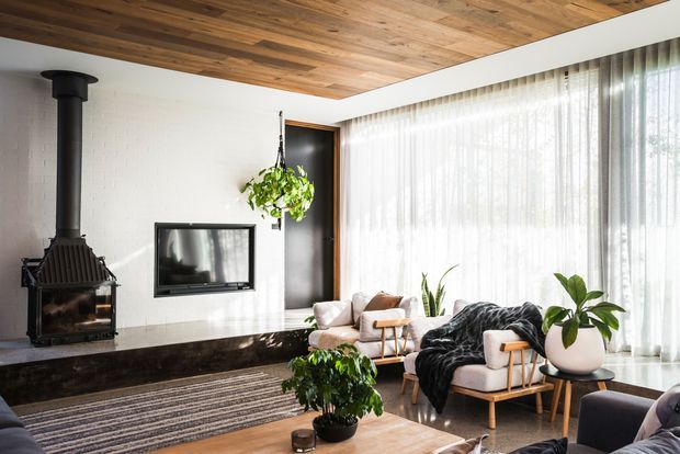 Bright contemporary living room with a black freestanding wood-burning fireplace, wall-mounted television, white-painted brick walls, full-height sheer curtains, and a warm timber-lined ceiling. Pale upholstered lounge chairs, indoor plants, and a timber coffee table create a relaxed, natural interior with strong daylight.