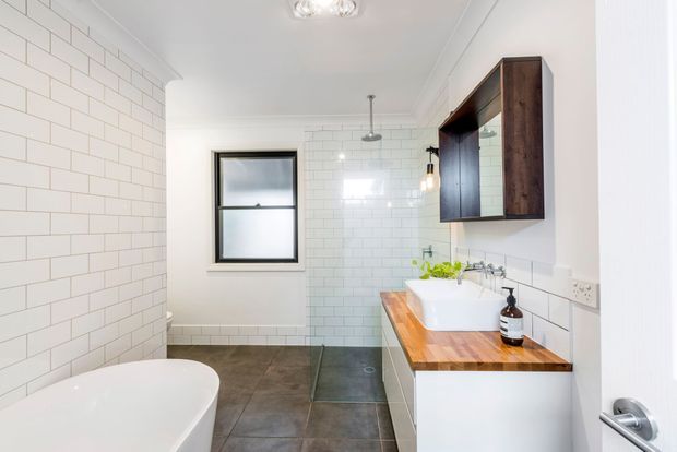 Bright contemporary bathroom with white subway-tile walls, dark grey floor tiles, a freestanding white bathtub, and a timber vanity with a white basin and dark wood wall cabinetry. Black-framed window and black fixtures provide contrast, while a glass shower enclosure is visible at the rear.