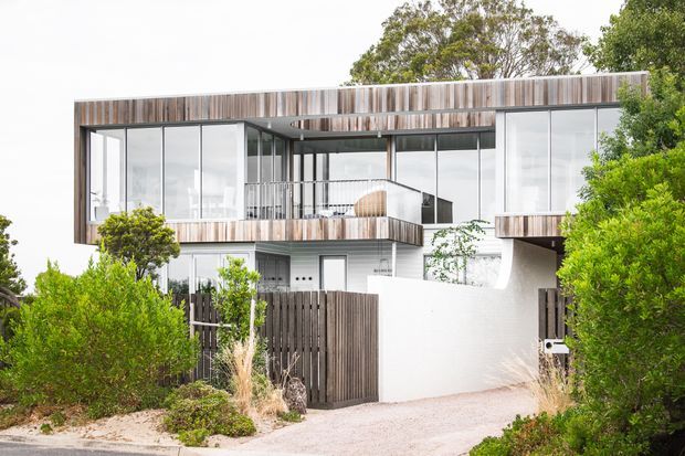 Contemporary multi-level residential facade with extensive floor-to-ceiling glazing, vertical timber screening and cladding, white rendered walls, and projecting balcony forms. The house is set behind landscaped planting with a timber privacy fence and driveway visible in the foreground.