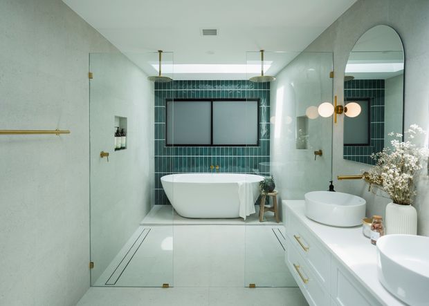 A bright, refined contemporary bathroom with a freestanding white bathtub behind clear glass shower panels, a double white vessel-sink vanity, pale terrazzo-like wall and floor surfaces, and a dark green vertical-tile feature wall. Brass fixtures, framed mirrors, rounded arch detailing, and globe wall lights add warm decorative accents.