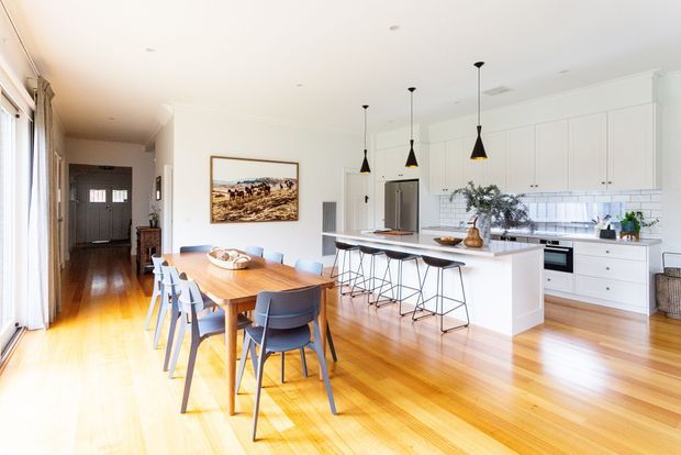 Bright open-plan kitchen and dining area with polished warm timber flooring, white shaker-style cabinetry, a large island with a white benchtop, tiled splashback, pendant lights, and a timber dining table with contemporary blue-grey chairs. A hallway and entry door are visible in the background.