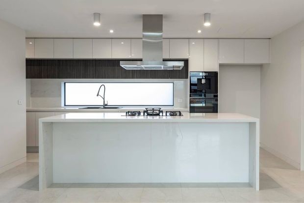 A bright, minimalist contemporary kitchen viewed straight on, dominated by a large white island with an integrated gas cooktop and sink. Full-height white cabinetry lines the rear wall, with a dark timber-toned horizontal feature band, pale stone-look surfaces, a stainless-steel rangehood, and built-in black appliances. Recessed and pendant-style ceiling lights provide even illumination.