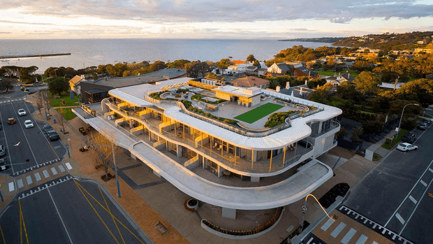 Aerial oblique view of a large contemporary multi-storey building with a rounded, highly glazed facade and sweeping white roofline. The rooftop contains landscaped terraces, planted areas, and a rectangular green-roof section. The building occupies a prominent urban corner beside marked streets and pedestrian crossings, with a coastal suburban landscape and water visible in the distance.