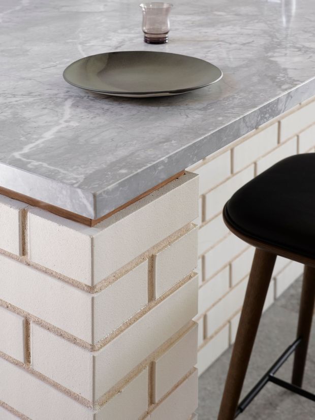 Close-up detail of a kitchen island or breakfast bar with a grey veined stone benchtop, white textured brick or blockwork base, and dark timber-and-black bar stool. A ceramic plate and small glass sit on the counter, highlighting the material palette and joinery detail.