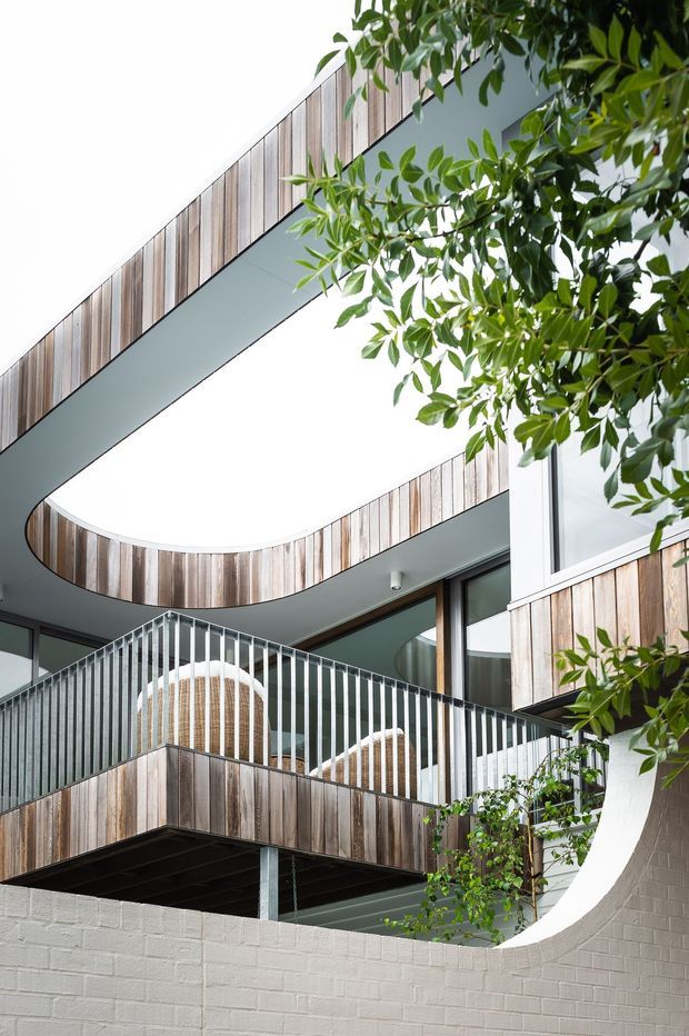 Exterior architectural photograph of a contemporary multi-level residential building focused on layered balconies, curved balcony edges, vertical white metal balustrades, timber-clad fascia and soffits, large glazing, and a light rendered brick boundary wall. Mature foliage partially frames the building.