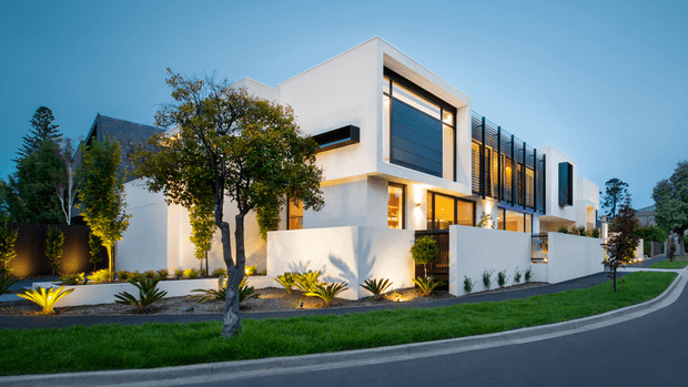 Contemporary multi-level white-rendered residence viewed from the street at dusk. The facade features strong rectilinear volumes, broad dark-framed glazing, projecting box forms, vertical screening, warm interior lighting, landscaped planting beds, and a curved paved street frontage.
