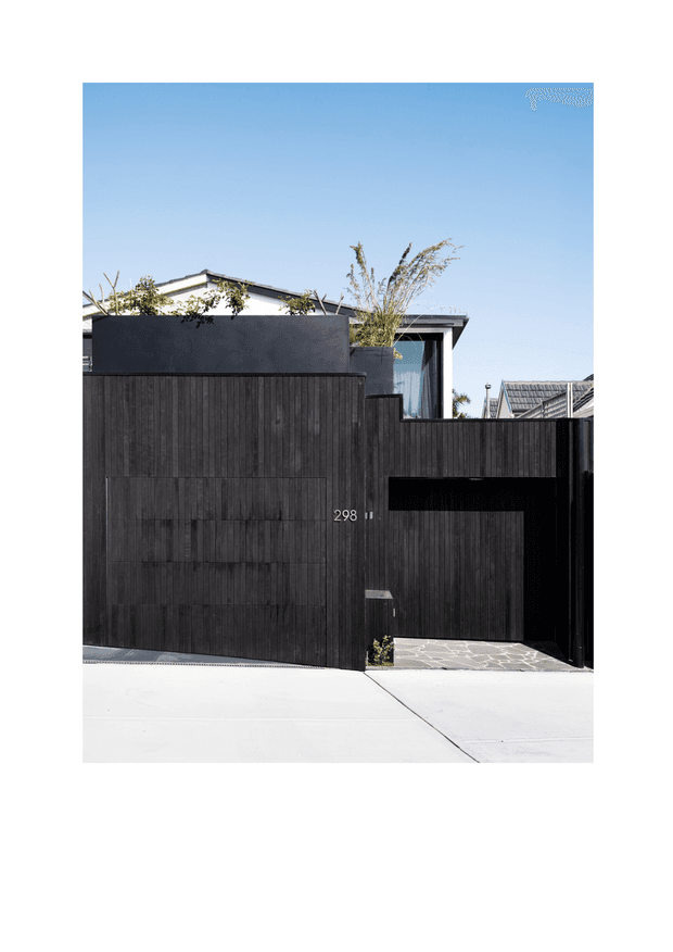 Street-facing view of a contemporary residential entrance with a dark vertical timber-clad boundary wall and matching pedestrian and garage gates. A minimalist flat-roofed house is partially visible behind the wall, with a narrow upper-level glazed opening, planted screening, and light stone paving at the entry. The image is presented against a white margin.