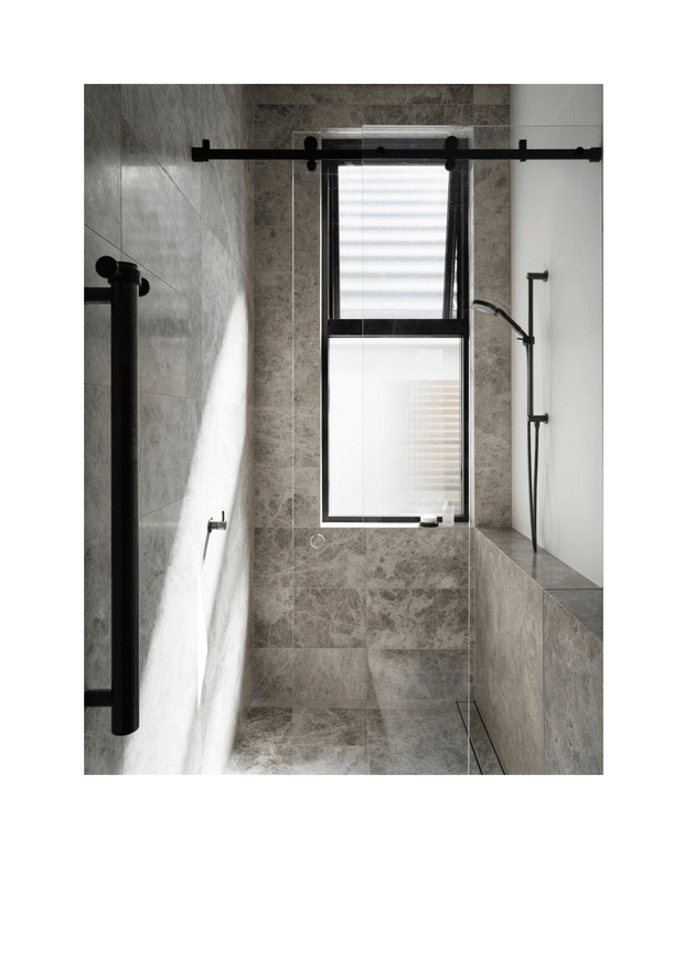 A contemporary walk-in bathroom shower lined with large grey stone-look tiles. The space features matte black exposed shower hardware, a black-framed window with a roller blind, a frameless glass shower screen, and a built-in tiled ledge. Natural light enters through the window, creating a calm, refined atmosphere.