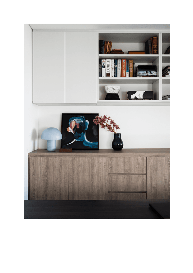 A clean, contemporary built-in storage wall featuring white overhead cabinets and open shelving displaying books and decor, above a wood-grain credenza with integrated drawers and doors. The styling includes a blue table lamp, artwork, and dark vase, creating a refined study or living area vignette.