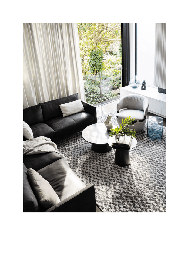 Bright, refined living room with a dark sectional sofa, sculptural round coffee tables, a curved upholstered armchair, textured grey patterned carpet, full-height sheer curtains, and tall black-framed glazing overlooking greenery. White built-in cabinetry and restrained decorative objects create a contemporary monochromatic interior.