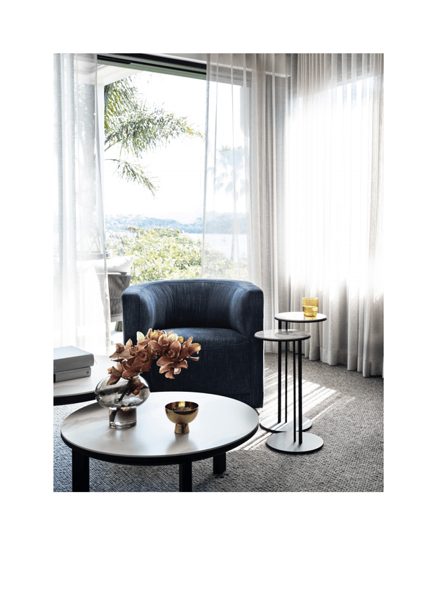 A refined living room seating area with a dark navy upholstered armchair, round black coffee table, slim nesting side tables, and floor-to-ceiling sheer curtains. Strong natural light enters through expansive glazing, with a leafy landscape and water view visible beyond. The room has a neutral textured carpet and a restrained, elegant furniture arrangement.