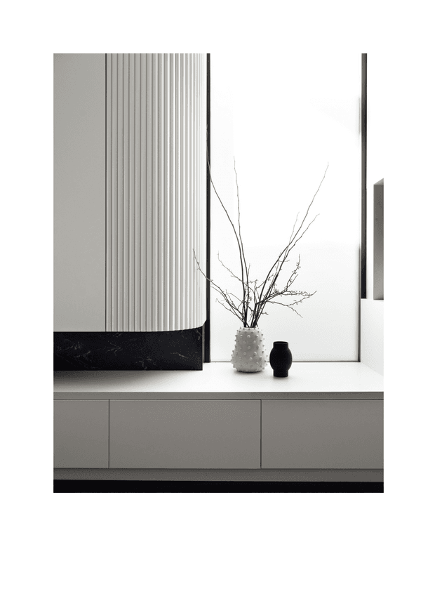 A minimalist interior vignette featuring a low, light-grey built-in cabinet with handleless doors, a dramatic black stone base or plinth, and a tall vertically fluted architectural panel. A bright recessed window or mirror-like opening forms the backdrop, with a small textured white vase containing bare branches and a black vessel placed on the cabinet. The composition is monochromatic, refined, and highly sculptural.