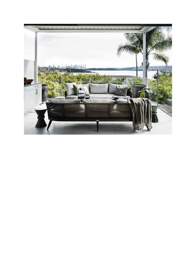 A bright, contemporary indoor-outdoor living area with a large upholstered sofa facing expansive glazing and a waterfront cityscape. The space includes a compact white built-in kitchenette or cabinet run at left, pale tiled flooring, slim dark-framed glazing, a covered flat-roof structure, potted palms, and neutral cushions and throws. The setting appears elevated with views across water toward a distant urban skyline.