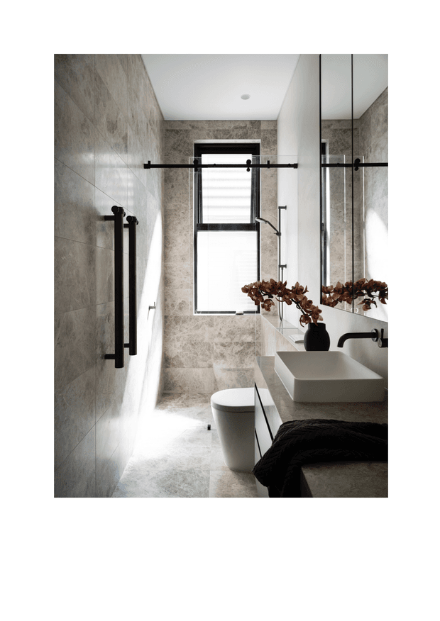 A narrow, contemporary bathroom with large grey-beige stone-look wall and floor tiles, a frameless glass shower enclosure, black shower hardware and towel rails, a wall-hung vanity with a white vessel basin, large mirrored cabinetry, and a compact white toilet. Natural light enters through a high window with a black frame and roller blind. A dark towel and dried botanical arrangement add contrast.