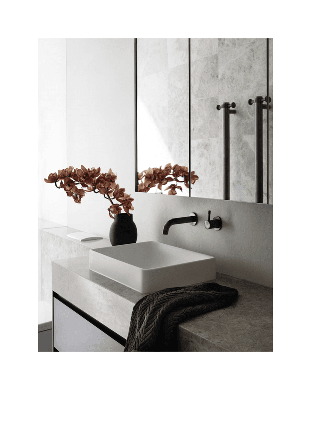 A refined contemporary bathroom vanity with a rectangular vessel basin set on a pale stone benchtop, matte black wall-mounted tapware, and a dark integrated vanity drawer. Large mirrored panels reflect the textured light stone wall and tapware. A black ceramic vase with dried brown botanical stems and a folded dark textured towel add restrained decorative accents.