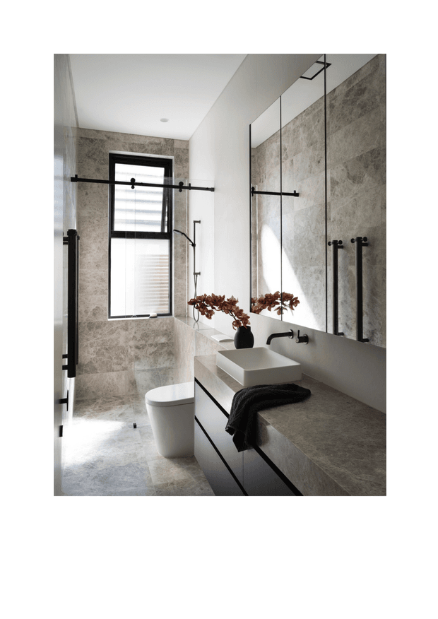 Contemporary bathroom with a walk-in shower enclosed by black-framed sliding glass doors, large-format grey stone-look wall and floor tiles, a floating dark vanity with a pale stone benchtop, vessel basin, wall-mounted black tapware, toilet, and full-width mirror. Natural light enters through a tall black-framed window, creating a refined, monochromatic interior.