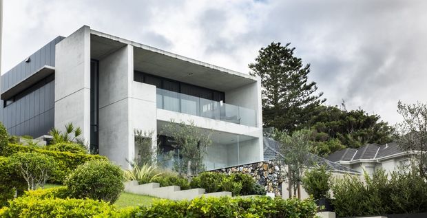 Exterior view of a contemporary multi-level residence set into a landscaped suburban site. The building features strong rectilinear concrete frames, expansive glazing, recessed balconies, dark metal wall cladding, and a flat roof, with a neighbouring pitched-roof house visible to the right.