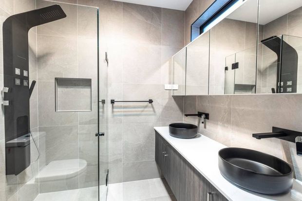 Contemporary bathroom featuring a double vanity with dark vessel basins, a white benchtop, vertically textured dark cabinetry, large grey-beige wall and floor tiles, a frameless glass shower enclosure, black shower fixtures, and a large mirrored wall.
