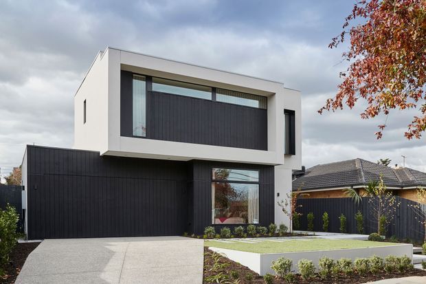 Contemporary two-storey residential house viewed from the street, with a crisp white rendered upper volume, dark vertical cladding, broad recessed upper-level glazing, and an integrated double garage. The landscaped front garden includes a paved driveway, raised lawn, low hedges, and dark boundary fencing. Neighbouring homes and autumn foliage are visible in the background.