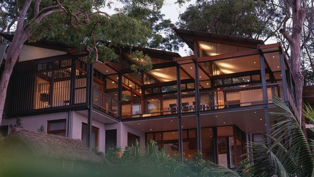 Exterior view of a multi-level contemporary home set among mature trees and landscaped planting. The house features extensive glazing, dark slender structural framing, elevated decks and balconies, warm interior lighting, and a series of angular sloping roof planes.