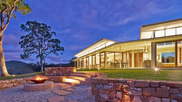 Twilight exterior view of a contemporary, low-profile residence set on a landscaped sloping site. The home features extensive floor-to-ceiling glazing, warm interior lighting, broad roof overhangs, and a combination of light rendered walls and timber-toned framed openings. In the foreground are layered dry-stone retaining walls, gravel landscaping, stepping stones, and a circular outdoor fire pit. Mountains and mature trees form the distant setting.
