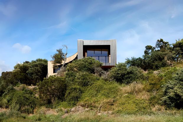 A modern elevated house partially concealed within dense coastal-looking vegetation. The visible facade features a rectilinear upper volume with vertical ribbed cladding or screening, deep window openings, and a sheltered balcony, with a curved or angled lower structure extending to the left. The image is an exterior architectural view set against a bright blue sky and landscaped bushy terrain.
