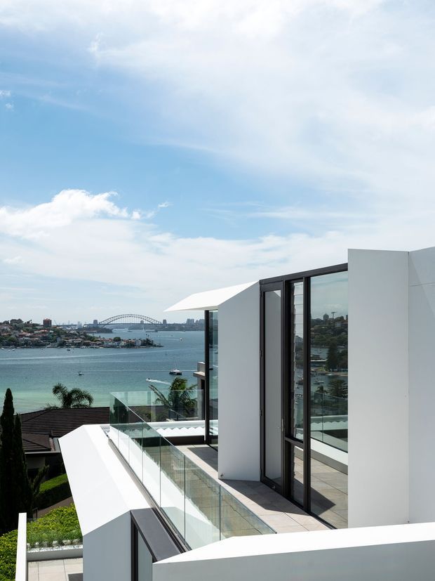 Contemporary white geometric residence viewed from an elevated exterior terrace, with expansive black-framed glazing, glass balustrades, tiled outdoor surfaces and a prominent harbour outlook featuring a bridge and surrounding suburbia.