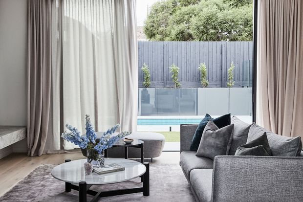 Refined contemporary living room with a large grey sectional sofa, round marble-topped coffee table, soft layered curtains, and neutral furnishings. Full-height glazing opens toward a private courtyard with a swimming pool and planted boundary.