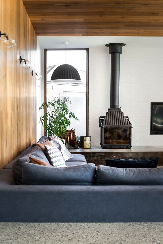 A warm, refined living room viewed from behind a large charcoal sectional sofa. The room features a dark timber-lined ceiling, vertical timber wall panelling, a white painted brick feature wall, a freestanding black wood-burning fireplace, and a large window. Globe wall lights, cushions, plants, and a television complete the space. A speckled terrazzo floor is visible in the foreground.