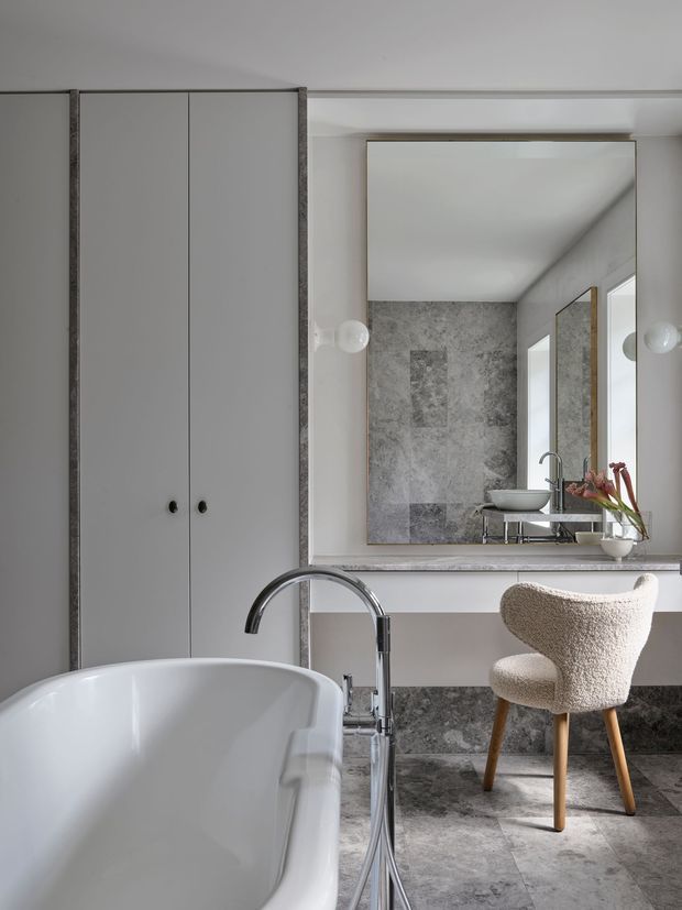 A refined contemporary bathroom featuring a freestanding white bathtub in the foreground, tall grey built-in cabinetry, a stone-clad vanity wall, large framed mirror, vessel basin, chrome tapware, and a textured upholstered stool. Grey stone flooring and wall finishes create a cohesive, muted palette, while globe wall lights and a small floral arrangement add detail.