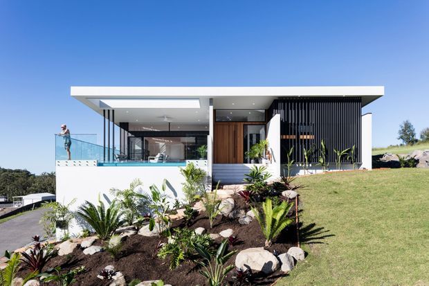 A contemporary single-storey residence set on a landscaped sloping site, featuring a broad flat roof, extensive glazing, white rendered walls, a timber entry door, and a dark vertical louvred screen. A planted garden bed with tropical and ornamental greenery occupies the foreground, while a glass-balustraded terrace is visible at the left.