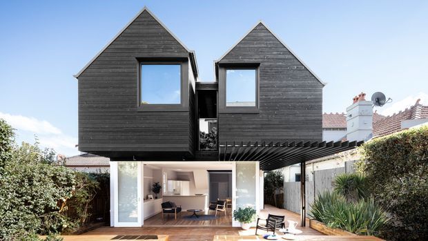 Contemporary two-storey residential extension with two dark timber-clad gabled volumes above a bright glazed living area. The lower level opens onto a paved courtyard or terrace, with a projecting dark pergola or canopy and established garden screening. A neighbouring traditional tiled roof and chimney are visible in the background.