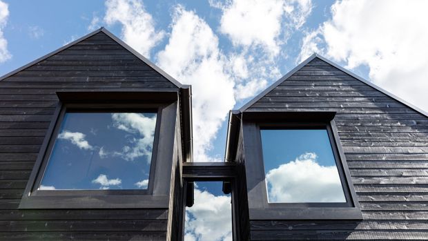 Close-up exterior view of two adjoining steep-gabled building forms clad in dark horizontal timber boards. Large projecting windows with dark frames reflect the bright blue sky and clouds, while a narrow glazed connection links the two volumes.