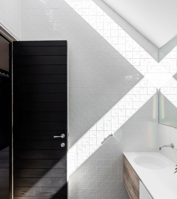 Bright contemporary bathroom with a black horizontal-panel door, white textured geometric wall tiles, and strong diagonal sunlight casting shadows across the wall. A white vanity with a light timber front, integrated basin, chrome tapware, and a large mirror are visible.