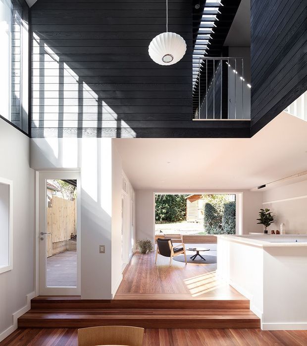Bright contemporary open-plan interior with a dramatic double-height void. Dark horizontal timber wall and ceiling panelling define the upper level and stair/landing, while white walls, a white kitchen island, warm timber flooring, and broad glazing create strong contrast. A sculptural pendant hangs in the void, with a dining and living area visible beyond the stepped floor transition.