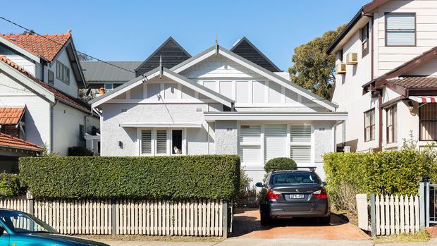 Street-facing view of a compact, light-coloured residential house with a prominent layered gabled roof, white rendered facade, external shutters, and a small front garden enclosed by trimmed hedges and a picket fence. A driveway and parked car sit in front, with neighbouring homes closely adjoining the property.
