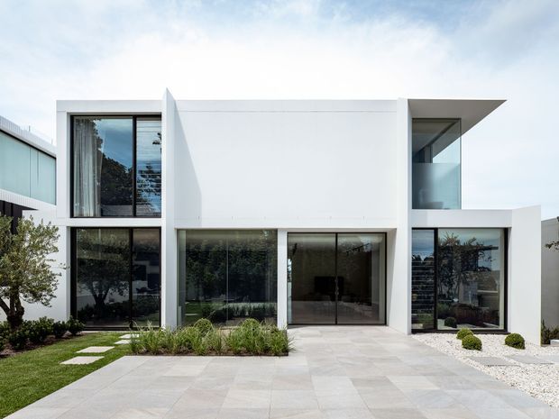 A contemporary two-storey white geometric residence with a boxy flat-roofed form, extensive dark-framed glazing, recessed balconies, and a broad pale-paved forecourt. The landscaped frontage includes clipped shrubs, ornamental grasses, stepping stones, and lawn.