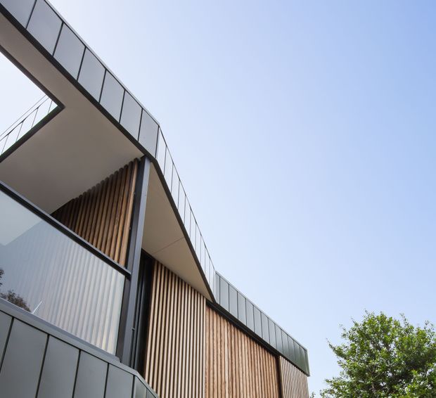 Upward-looking exterior view of a contemporary multi-level building with sweeping cantilevered roof or balcony forms, dark metal-framed glazing and balustrades, and vertical timber slat cladding. A tree and clear sky surround the sharply articulated facade.